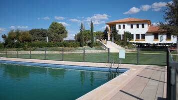 Piscina com superfície de água azul cristalina, ladeada por um deck pavimentado. Ao fundo, um edifício branco com telhado de telhas vermelhas e uma escadaria de pedra que leva a uma porta.