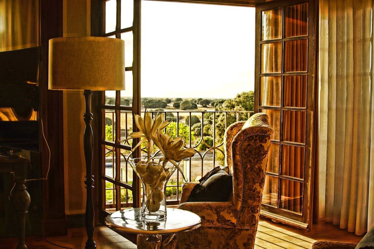 Une pièce avec un piano, une table sur laquelle se trouvent une lampe et un vase, et une vue sur la verdure par la fenêtre.