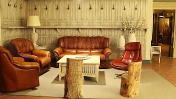 Een woonkamer met een bruine leren bank, twee fauteuils, een salontafel en een lamp. Er is houten wandpaneel en een vaas met takken op een tafel.