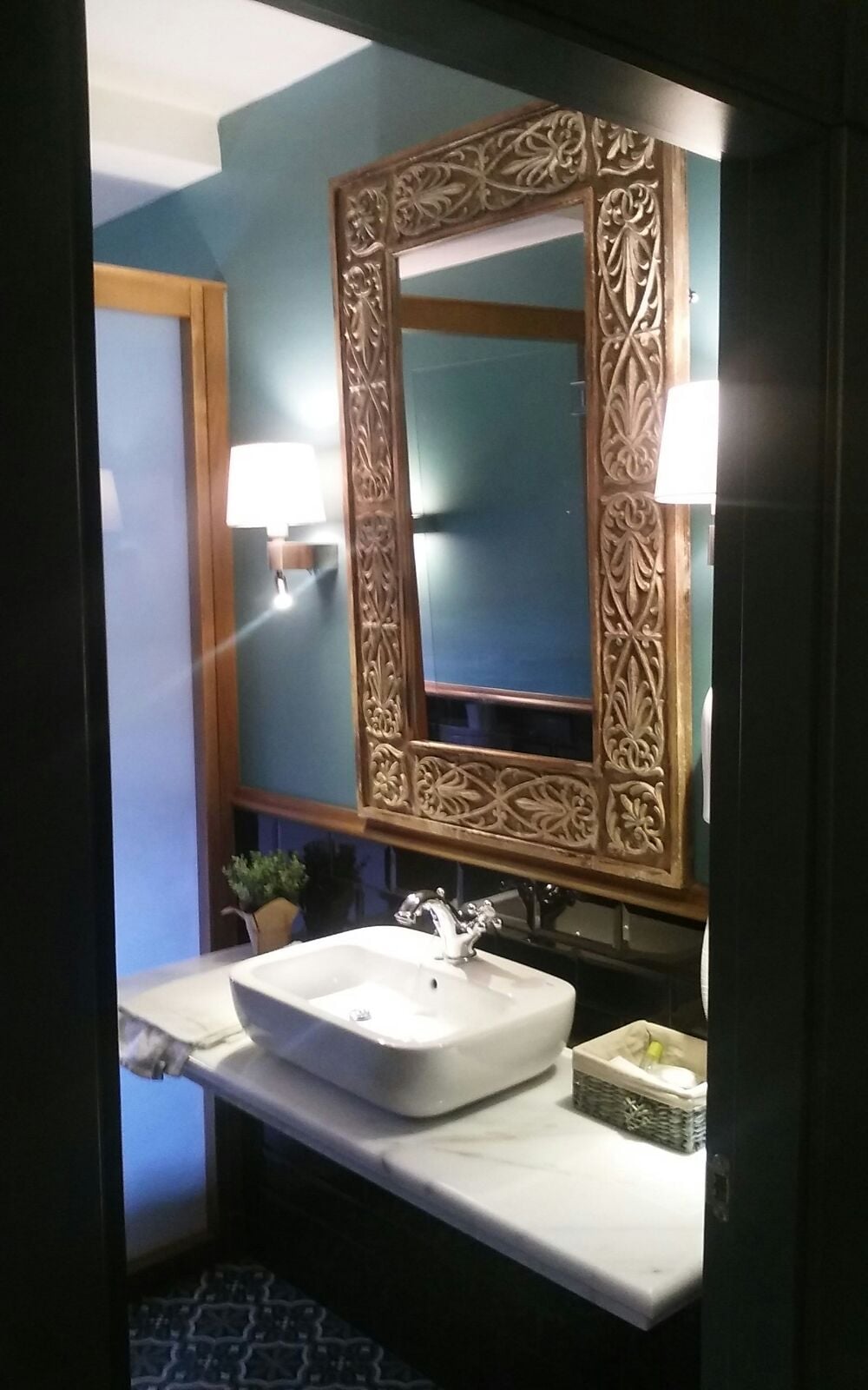 Une salle de bain avec un lavabo blanc, un miroir doté d’un cadre orné et une lampe fixée au mur.