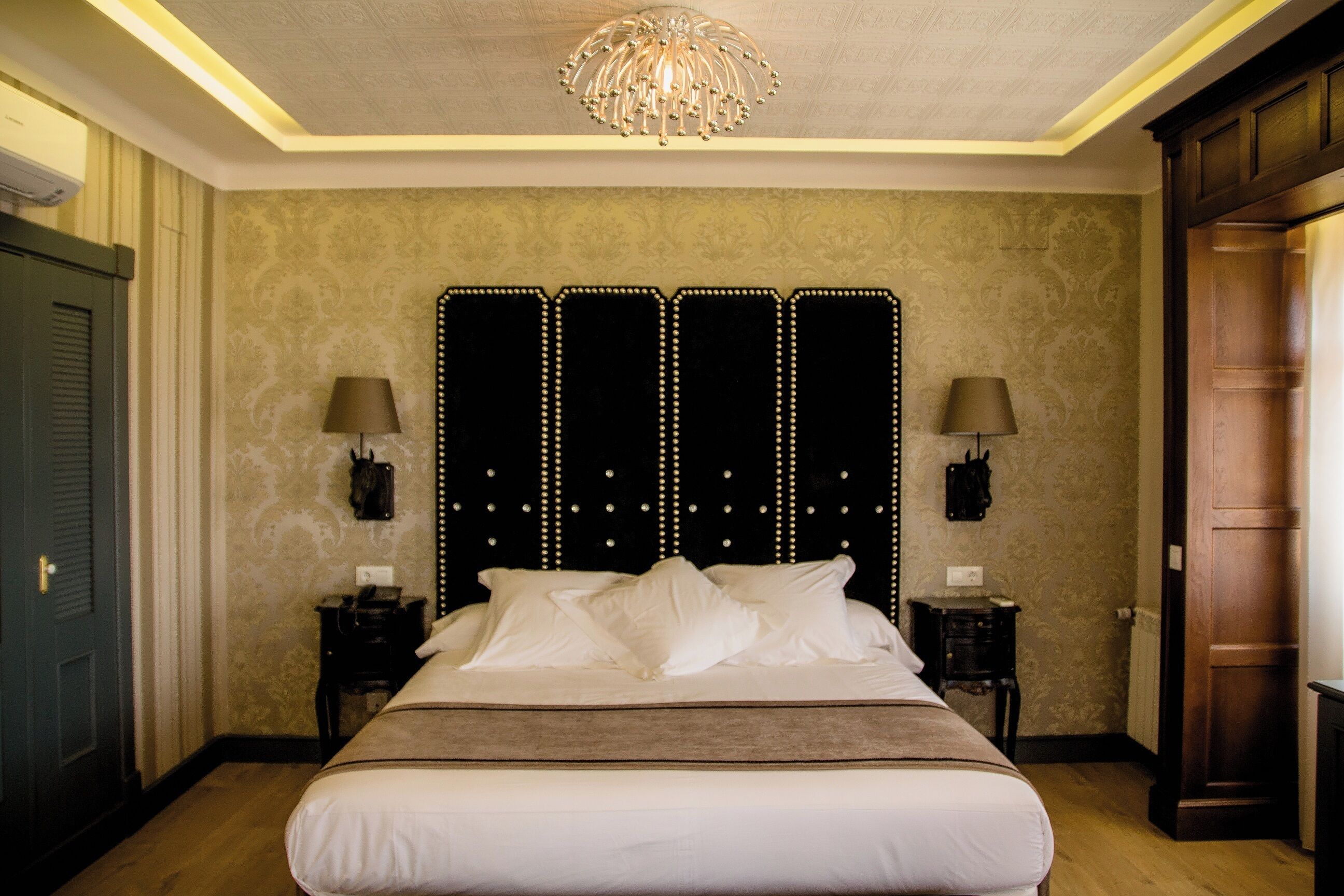 A hotel room with a large bed, two bedside tables, a chandelier, and a patterned wallpaper.