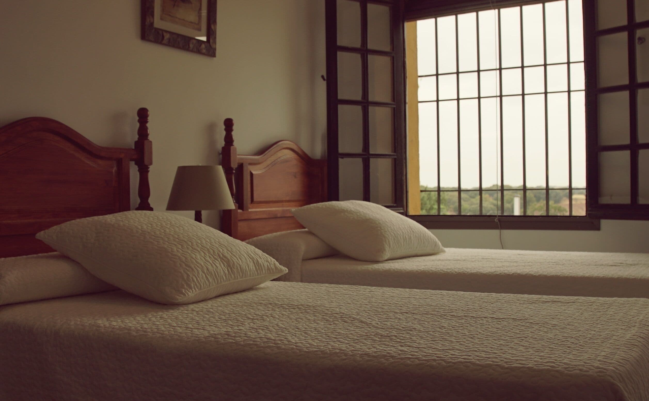 A room with two beds, a wooden headboard, a nightstand with a lamp, and a window with bars.