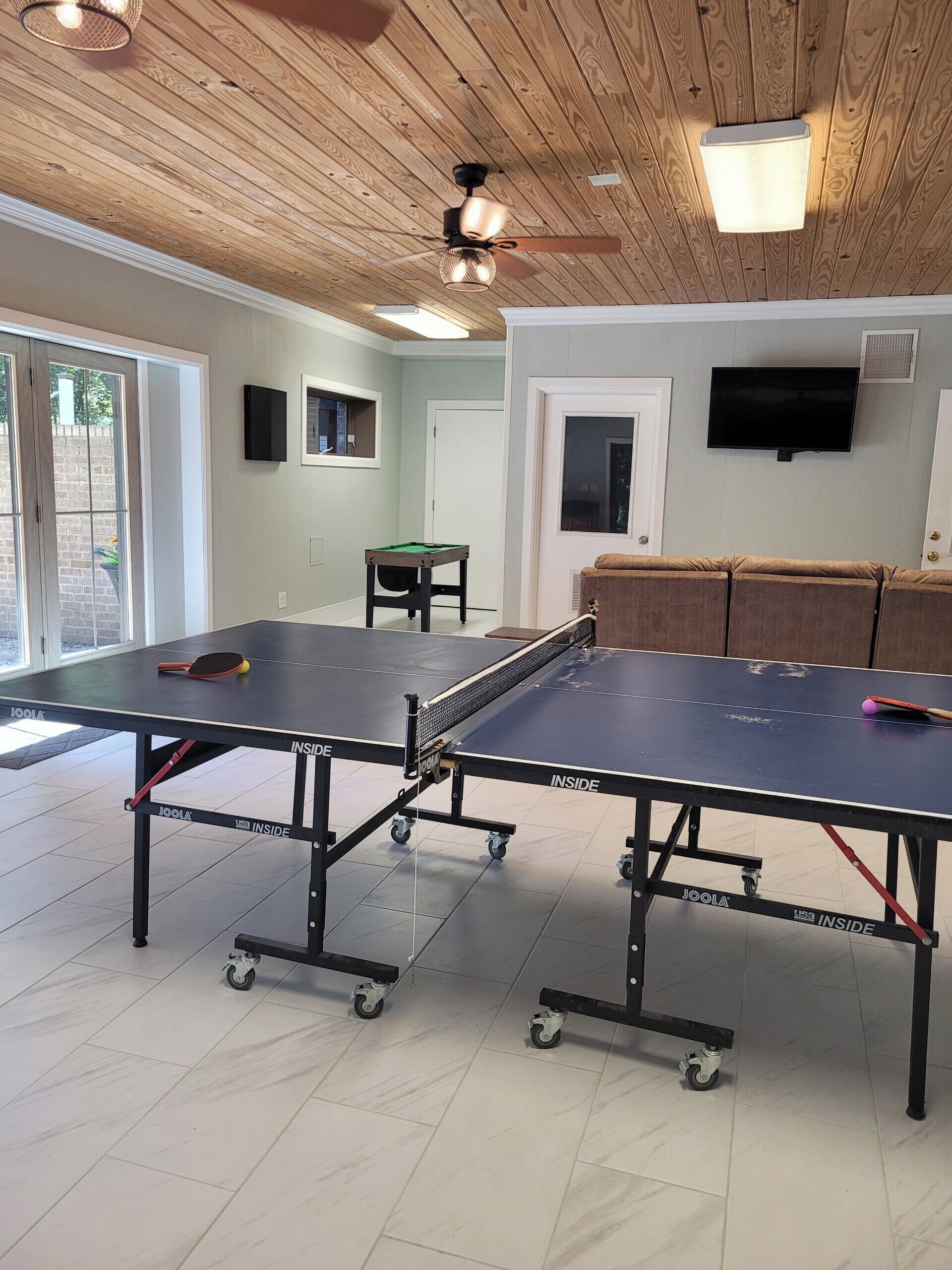 A room with a ping pong table, a sofa, a television, and a ceiling fan.