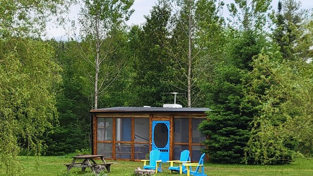 Een blokhut aan een meer, met een picknicktafel, twee blauwe stoelen en een klein terras.