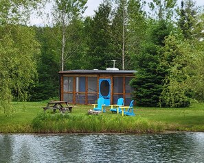 Une cabane au bord d’un lac, avec une table de pique-nique, deux chaises bleues et une petite terrasse.