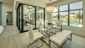 Un espace repas moderne avec une table en verre, des chaises blanches et un banc. Une grande fenêtre offre une vue sur une piscine et sur la verdure.