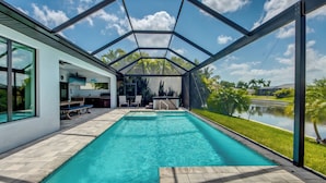 Une piscine extérieure moderne, dotée d’un patio fermé par des baies vitrées, d’un espace barbecue et offrant une vue sur un lac et des palmiers.