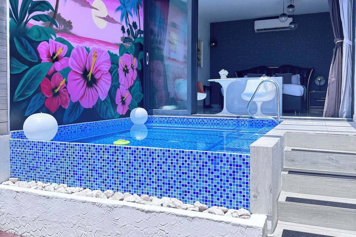 A modern pool area with a tropical mural, a glass-enclosed room, and a staircase leading to a bedroom.