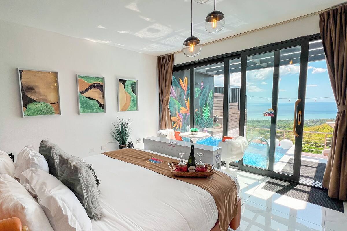 A modern bedroom with a large bed, a desk with a laptop, and a view of the ocean through large sliding doors.