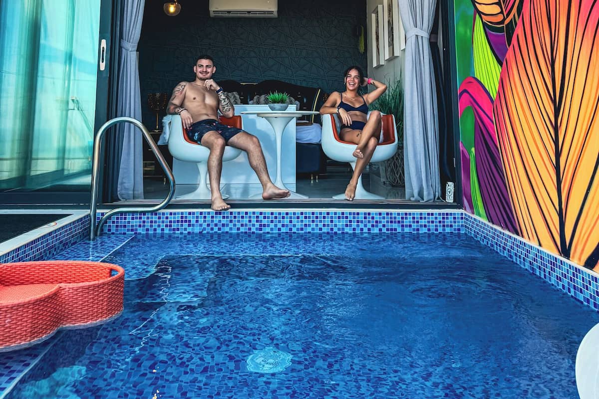 A modern indoor pool area with a glass ceiling, a colorful leaf-patterned wall, and a seating area with a table and chairs.