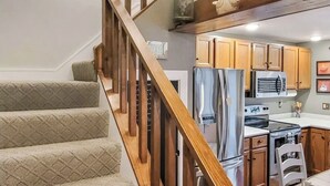 A staircase with wooden railings leading to a kitchen area with wooden cabinets, a refrigerator, an oven, and a dining table with chairs.