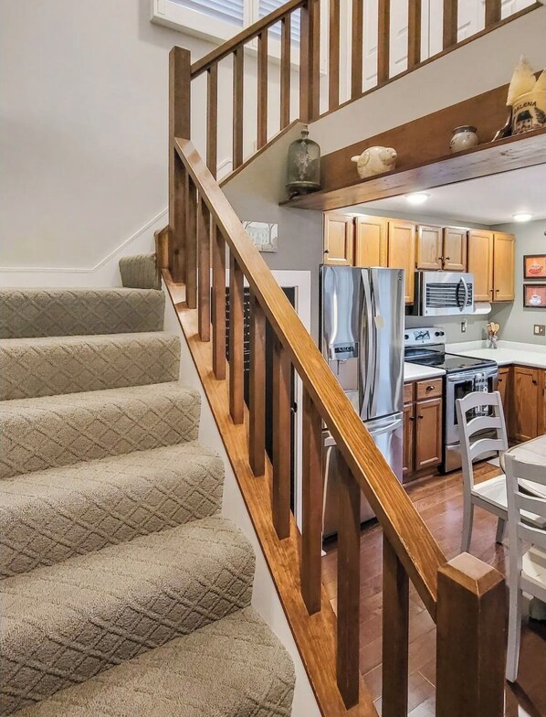 A staircase with wooden railings leading to a kitchen area with wooden cabinets, a refrigerator, an oven, and a dining table with chairs.