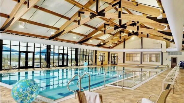 An indoor swimming pool with wooden beams, clear glass walls, and lounge chairs.
