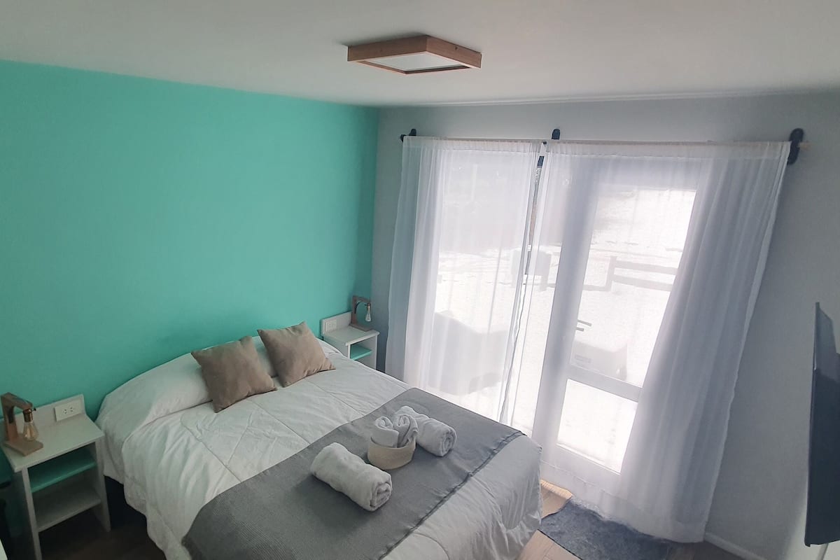 Un dormitorio con paredes verde mar, una cama con ropa de cama blanca y gris, una mesita de noche con lámpara y una ventana con cortinas de encaje.