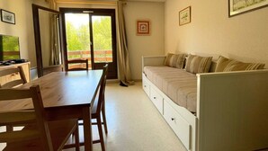 A room with a wooden dining table, a sofa bed, a television, and a balcony with curtains. - Studio le Coquelicot (L ESPEROU)