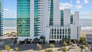 A modern hotel with a glass facade, located near a beach with palm trees and a clear sky. - Unforgettable Getaway 3BR (Myrtle Beach)