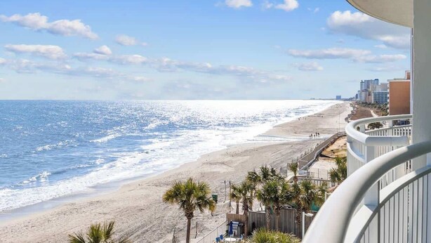 A beach view from a balcony with a railing, palm trees, and buildings in the background. - Unforgettable Getaway 3BR (Myrtle Beach)