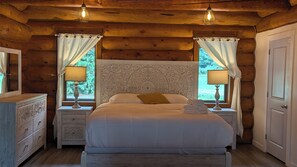 A wooden cabin bedroom with a large bed, two bedside tables with lamps, a dresser, and a door. - Opulent Oaks Chalet
 (Agassiz)