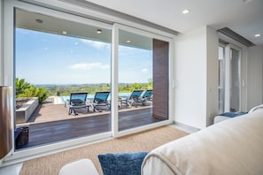 A modern living room with a large sliding glass door leading to an outdoor deck with lounge chairs and a pool.