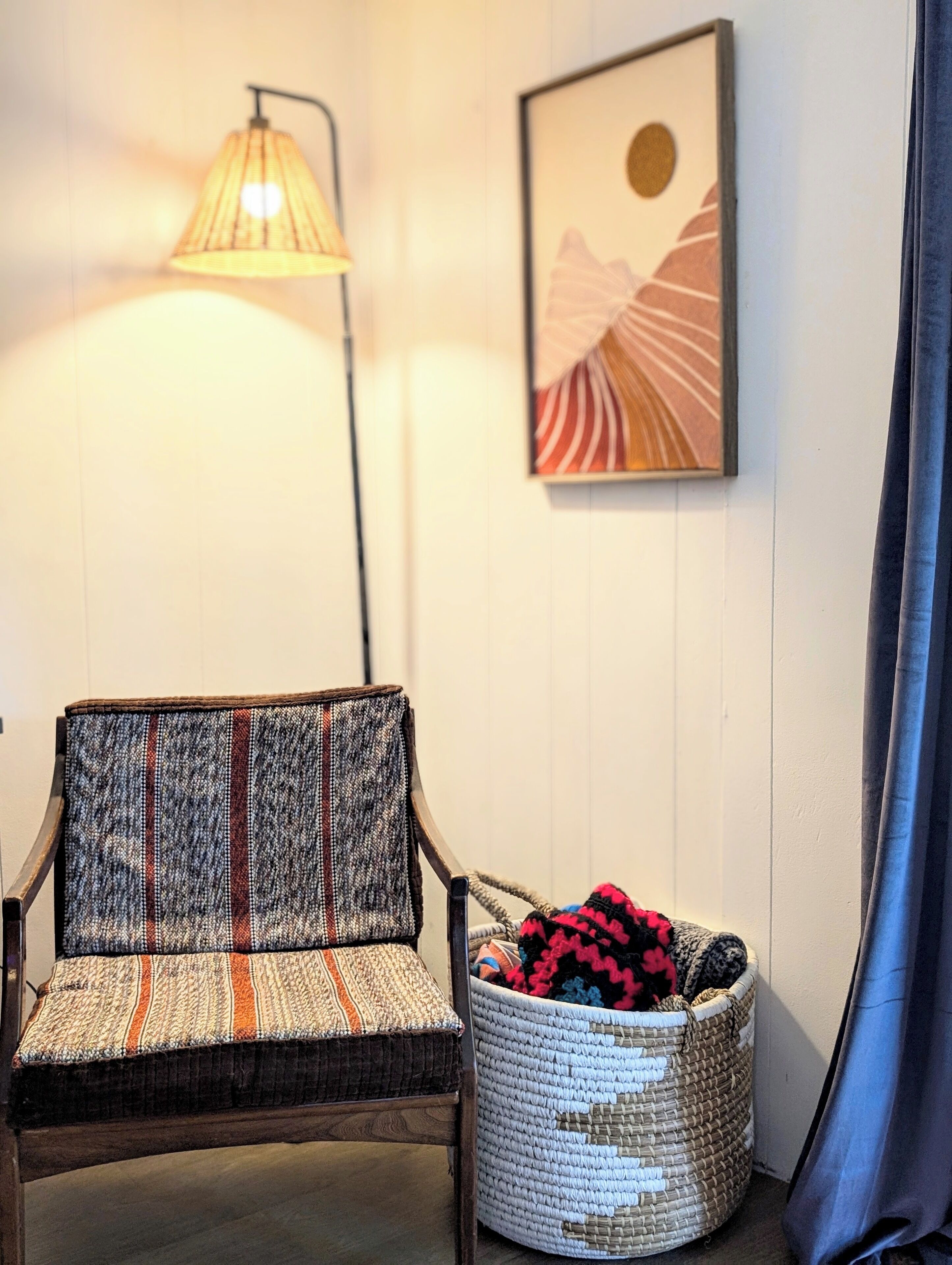 Una habitación con una silla de fibras naturales, una cesta tejida, una lámpara de pie y una obra de arte enmarcada en la pared.