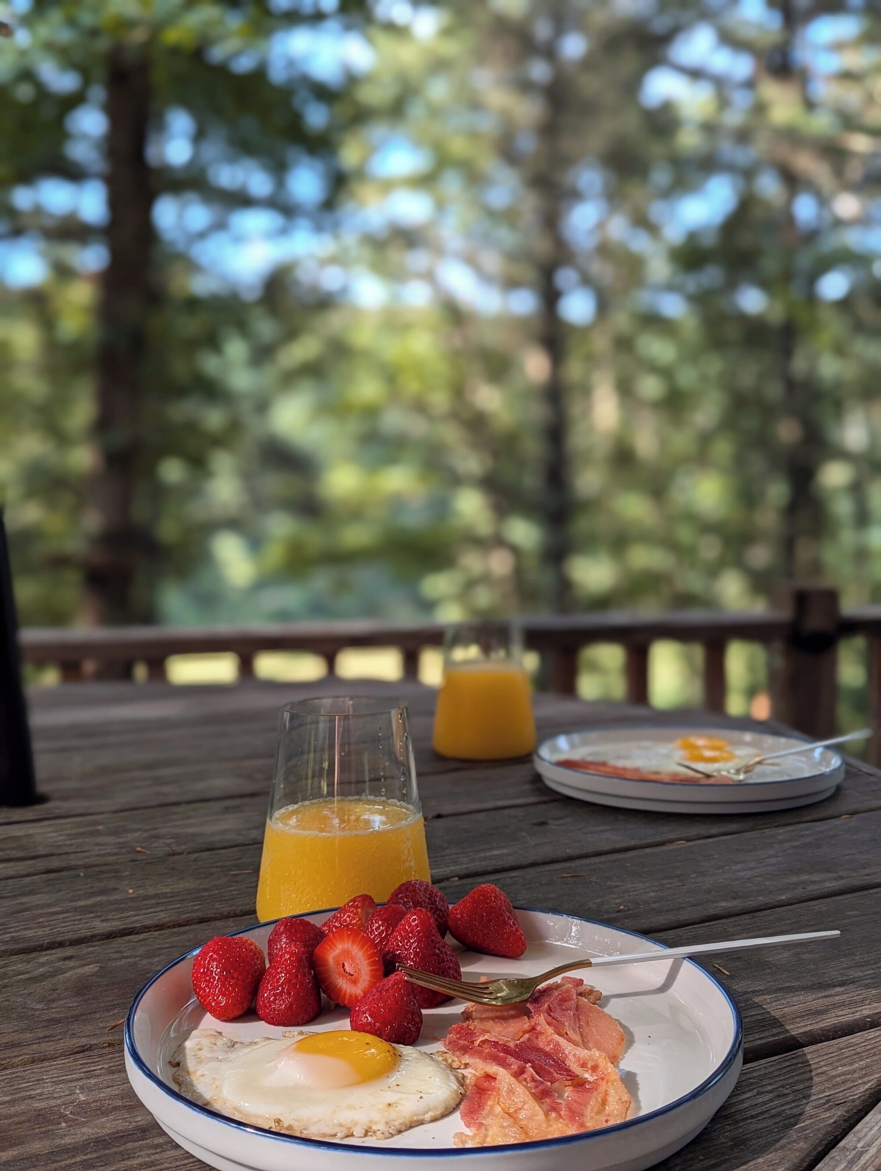 Une table de petit-déjeuner installée sur une terrasse en bois, avec une assiette d’œufs, de bacon, des fraises et un verre de jus d’orange.