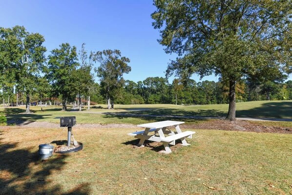 A picnic table and a trash bin in a park with trees and a clear sky.