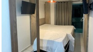 A bedroom with a bed, a TV mounted on the wall, a ceiling light, and a door leading to another room. - Israel Tambaú - APTO Luxuoso Beira Mar  (João Pessoa)