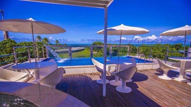 A pool area with a view of the ocean, surrounded by white umbrellas and lounge chairs. - Israel Tambaú - APTO Luxuoso Beira Mar  (João Pessoa)