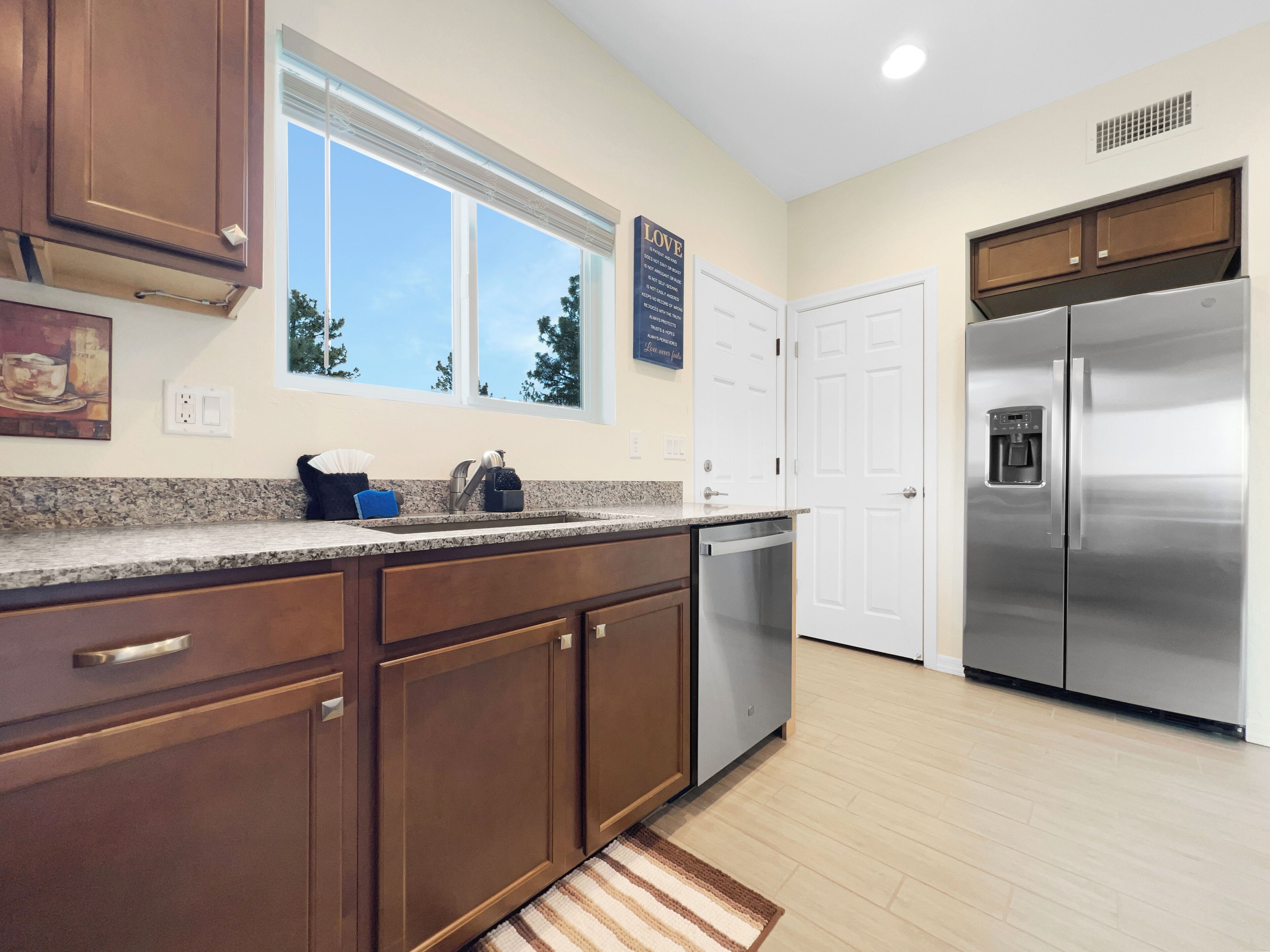 A modern kitchen with wooden cabinets, a stainless steel refrigerator, a dishwasher, and a window with a view of trees.