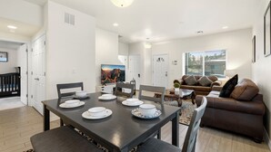 A modern living room with a brown leather sofa, a dining table set with white plates, and a large window with blinds.