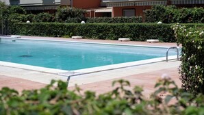 A rectangular swimming pool with a tiled deck, surrounded by neatly trimmed hedges and a row of white buildings in the background. - Terrazza sul Mare - Appartamento (Mascali)