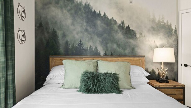 Un dormitorio con cama, almohadas, una mesita de noche, una lámpara y un mural en la pared con un bosque.