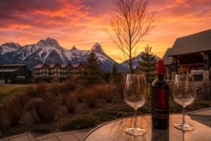 Ein Tisch mit einer Weinflasche und zwei Gläsern, vor einer Berglandschaft und einem Sonnenuntergang.
