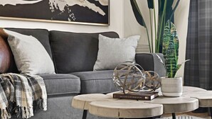 A living room with a grey sofa, a coffee table with decorative items, a large plant, and a framed artwork on the wall. - ★Mountain Haven★Pool&HotTub☆King Bed☆AC☆BBQ/Patio! (Canmore)