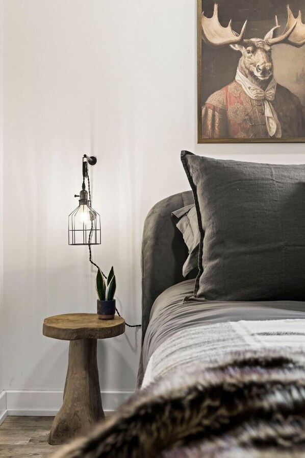 A bedroom with a bed, a small wooden side table, a potted plant, and a wall-mounted light fixture.