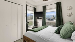 A bedroom with a bed, a chair, a desk, a window with curtains, and a view of the outside. - ★Mountain Haven★Pool&HotTub☆King Bed☆AC☆BBQ/Patio! (Canmore)