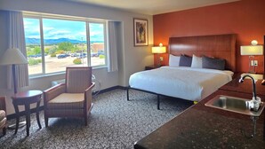 A hotel room with a bed, a chair, a small table, a desk with a sink, and a window with a view of a parking lot and mountains. - Olympia Inn  (Colorado Springs)