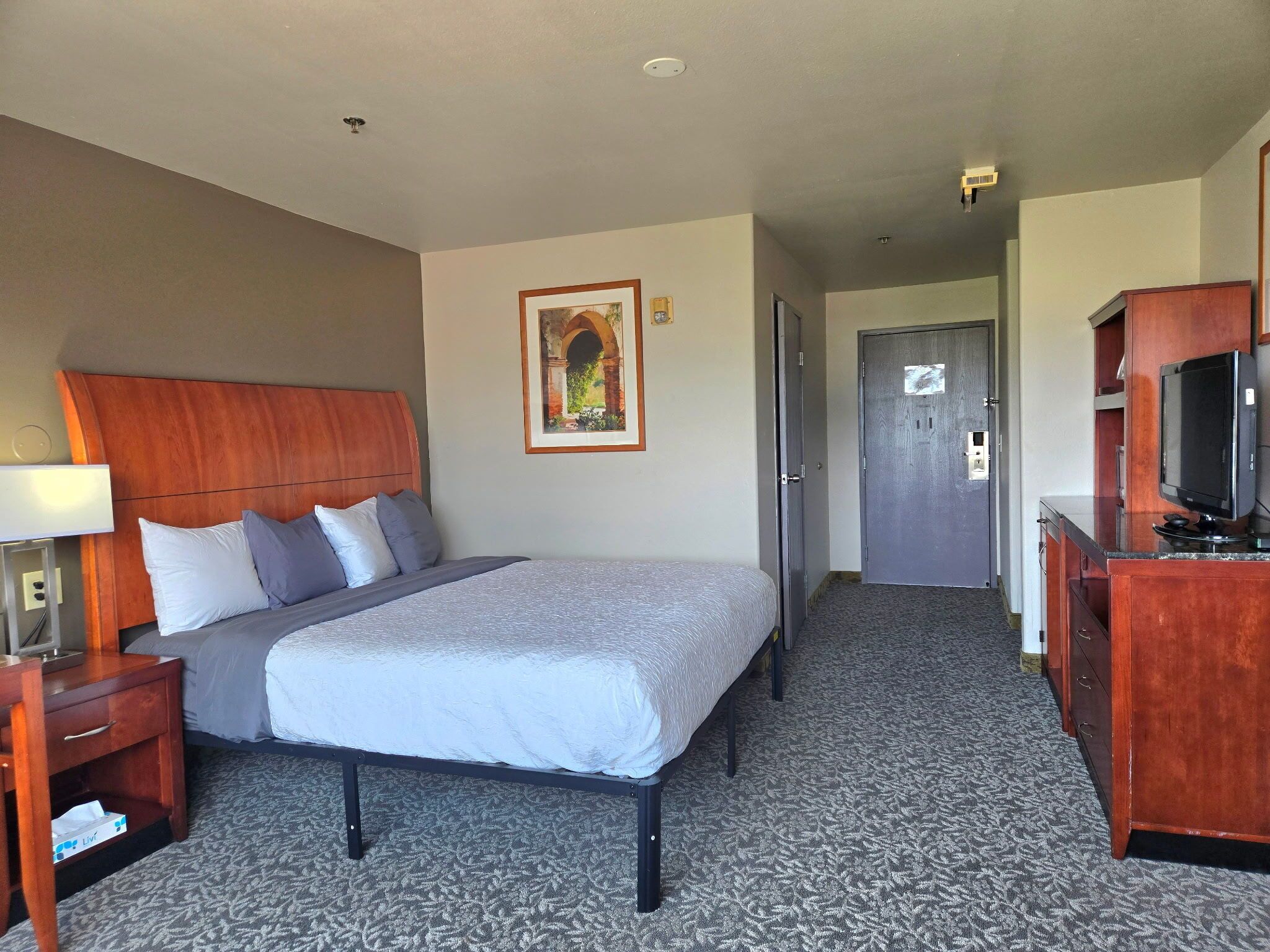 A hotel room with a bed, a desk, a TV, and a door leading to another room.