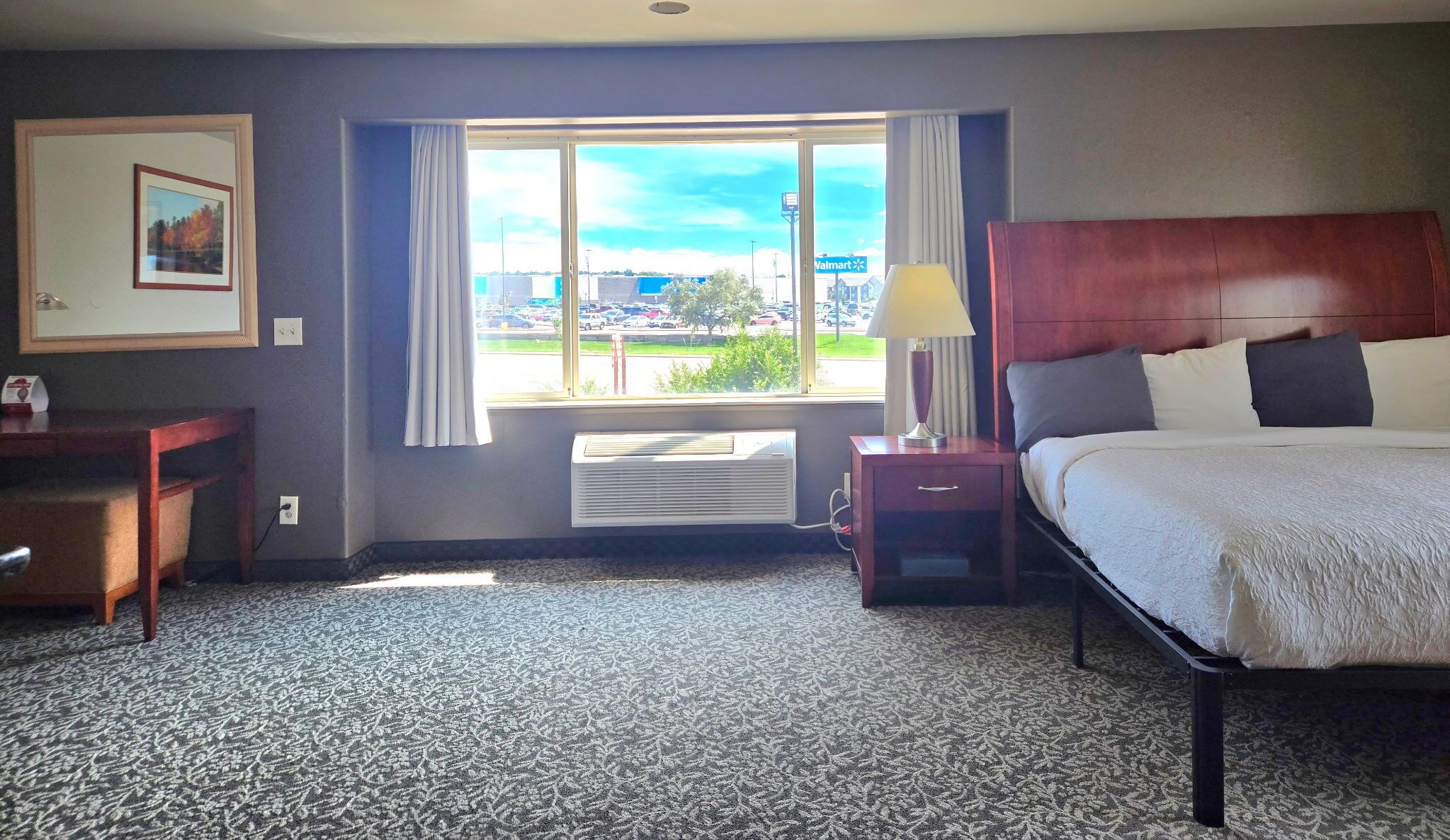 A hotel room with a bed, a desk, a chair, a window with a view of a parking lot and a Walmart store, and a framed picture on the wall.