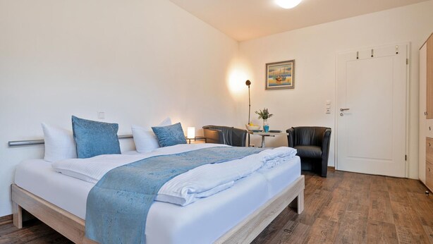 Un lit bien fait, recouvert d’un jeté bleu, une table de chevet avec une lampe, une chaise et une porte dans une pièce dont le sol est en bois.