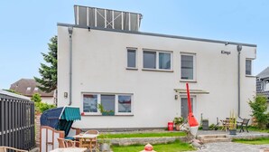 Un bâtiment moderne à deux étages, doté d’un toit plat, d’un espace extérieur abrité avec des tables et des chaises, et d’un petit jardin avec un chemin menant à l’entrée.