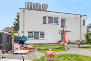 A modern two-story building with a flat roof, a covered outdoor seating area with tables and chairs, and a small garden with a pathway leading to the entrance. - Ferienwohnung 02 - Kimpi (Koserow)