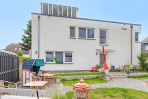 A modern two-story building with a flat roof, a covered outdoor seating area with tables and chairs, and a small garden with a pathway leading to the entrance. - Ferienwohnung 02 - Kimpi (Koserow)