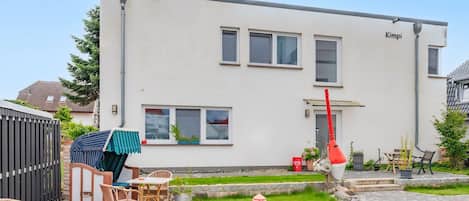Un bâtiment moderne à deux étages, doté d’un toit plat, d’un espace extérieur abrité avec des tables et des chaises, et d’un petit jardin avec un chemin menant à l’entrée.