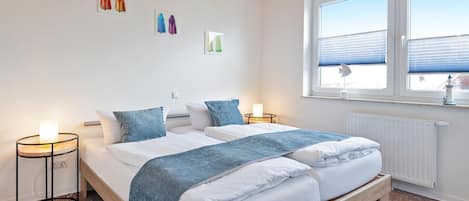 Une chambre à coucher avec un lit double, deux tables de chevet, une fenêtre avec des stores et des œuvres d’art accrochées au mur.