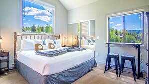 A bedroom with a large bed, two windows with views of greenery, a desk with a chair, and a nightstand with a lamp. - Modern Golf View Condo + Resort Amenities (Sun Valley)