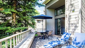 A wooden deck with a table and chairs, a blue umbrella, and a view of a house with a sliding glass door. - Modern Golf View Condo + Resort Amenities (Sun Valley)