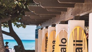 A row of surfboards with 'Hawaii' written on them, placed under a structure with a view of the ocean. - Beachside 2BR condo w/ lanais & free parking (Honolulu)