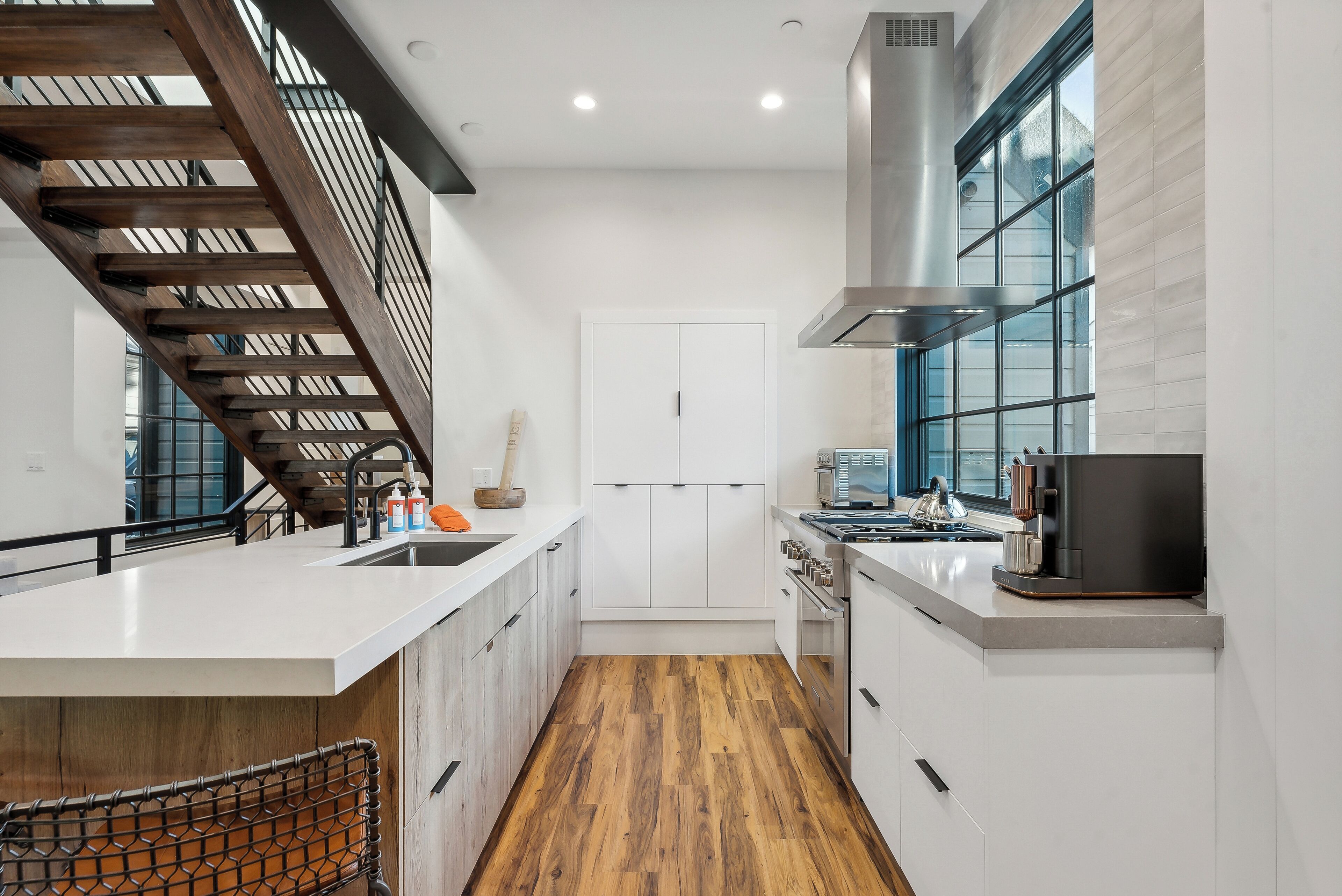 A modern kitchen with a wooden staircase, white cabinetry, and a large window.