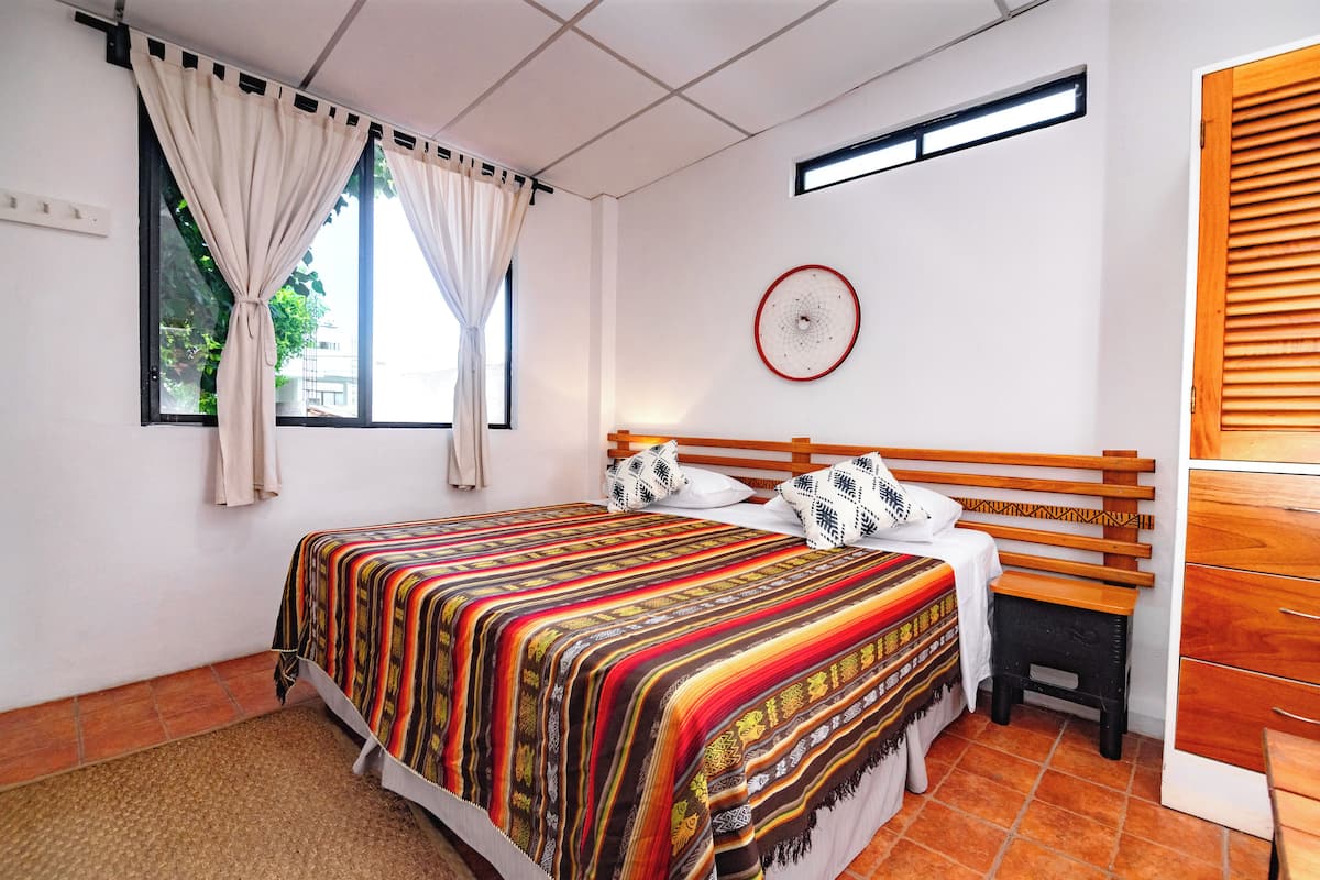 A bedroom with a bed, a nightstand, a wooden bench, a window with curtains, a wall clock, and a radiator.